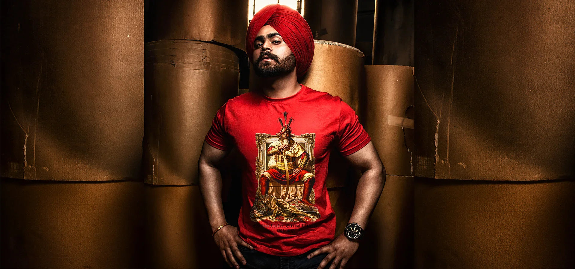Man in red turban and bold Punjabi graphic streetwear t-shirt standing among large brown rolls.